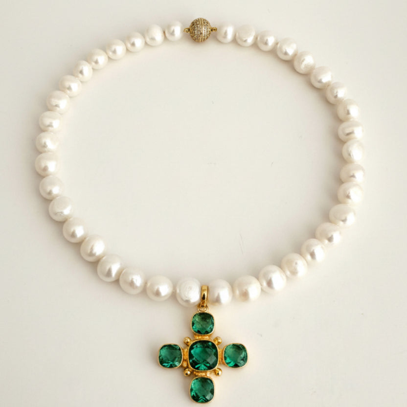 A white pearl necklace with a gold cross pendant featuring green gemstones, displayed on a light cream background.