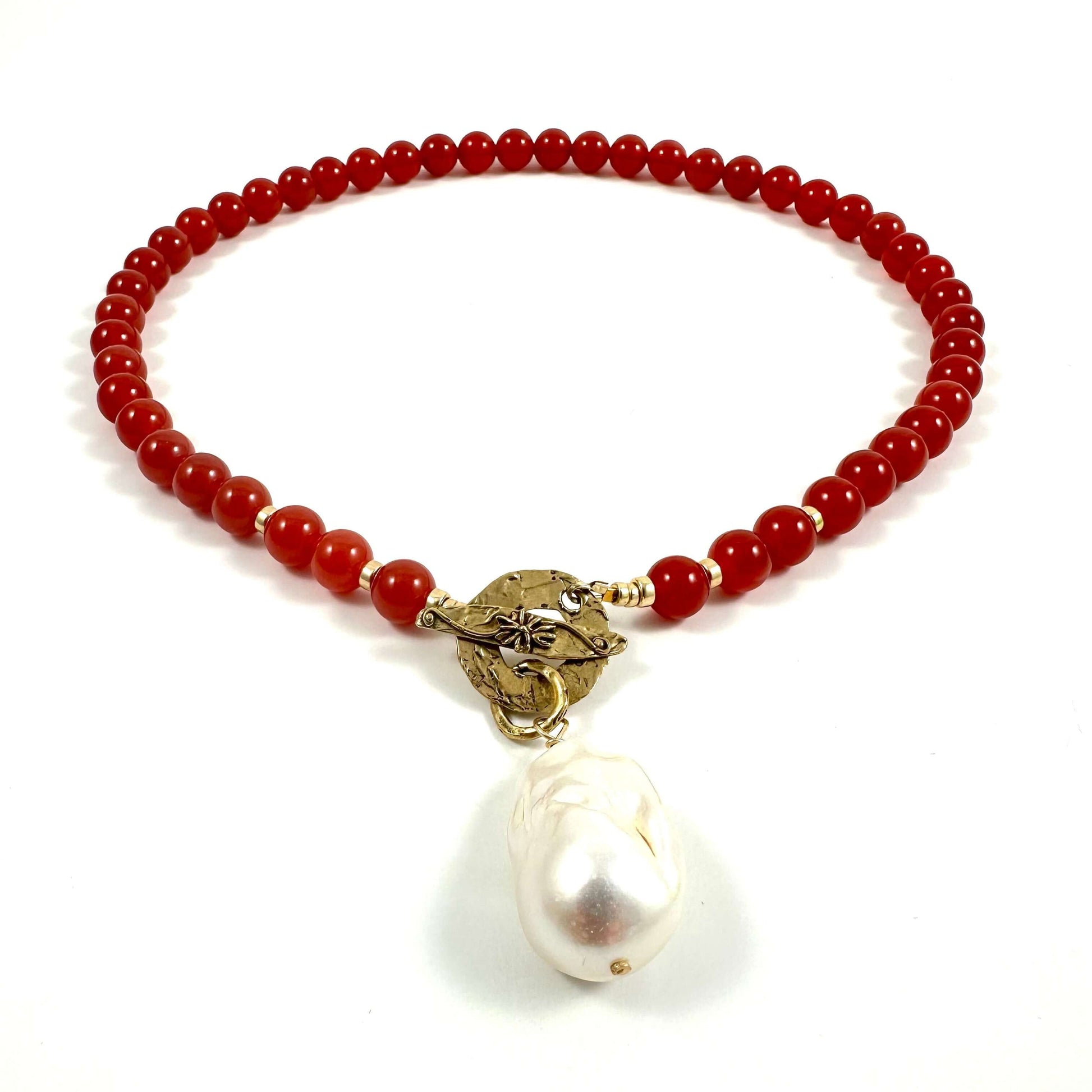 A beaded necklace featuring round red beads, a textured gold-tone metal connector, and a large, irregular white baroque pearl pendant, displayed on a clean white background.