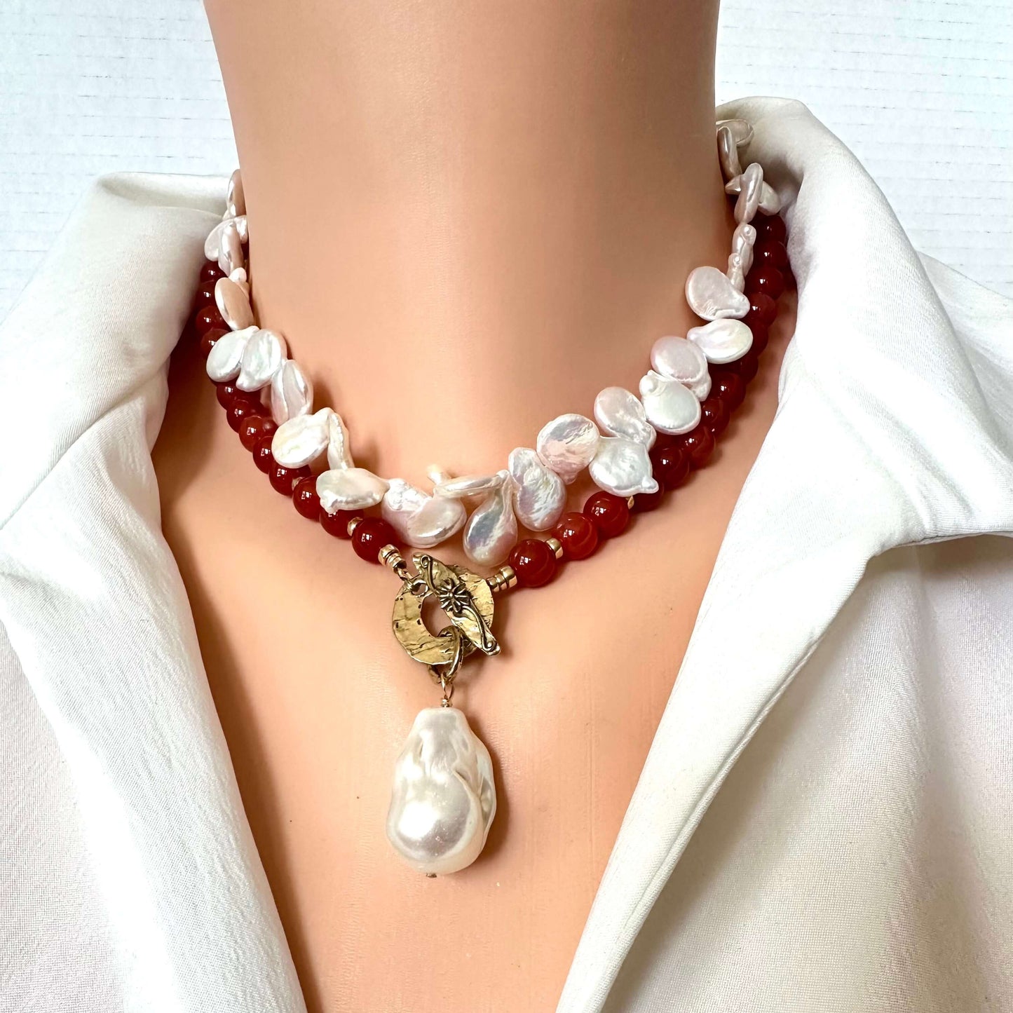 A close-up of a double-strand necklace worn on a mannequin torso. The necklace features dark red translucent beads paired with irregular white baroque pearls and a central gold-tone clasp holding a larger white pearl pendant.