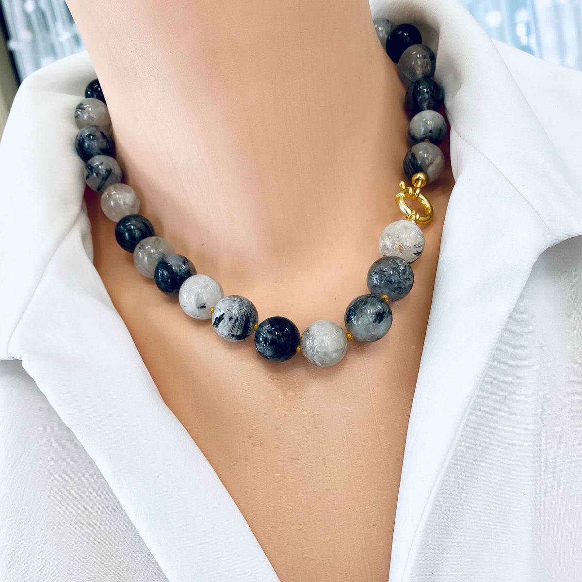 A close-up of a beaded necklace worn on a model's neck, featuring round gray, black, and white marbled beads with a small gold toggle clasp, styled with a white collared shirt.