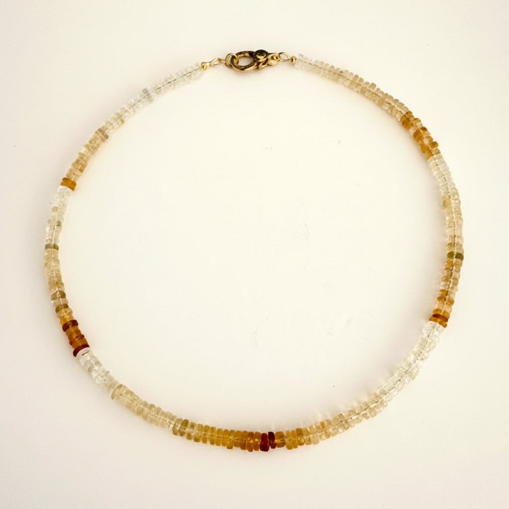 A beaded necklace made of small translucent and opaque beads in white, clear, and warm amber-brown tones, with a dark metal clasp, laid flat on a light off-white background.