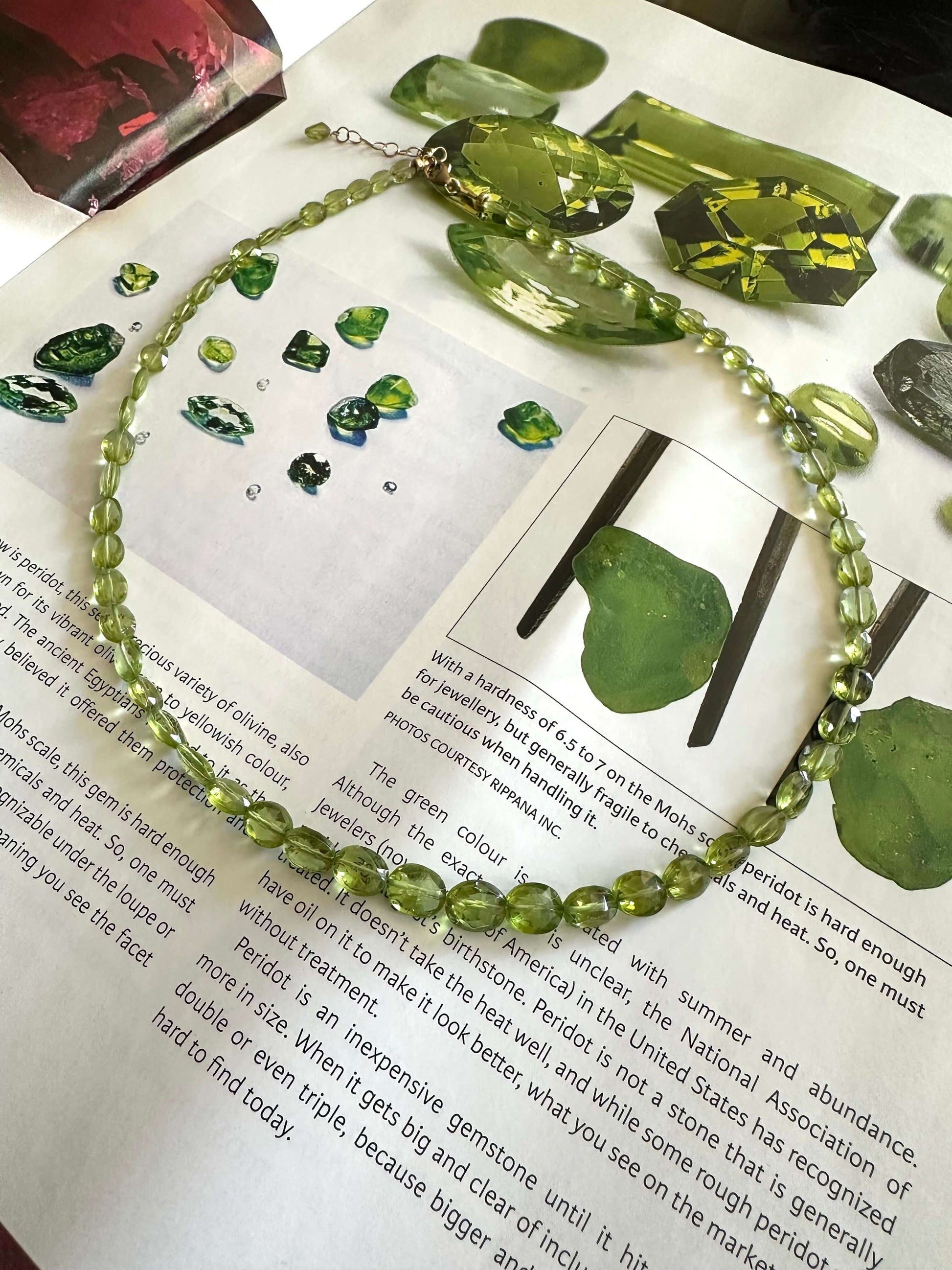 A green gemstone beaded necklace arranged on top of printed pages, surrounded by polished and rough green gemstones.
