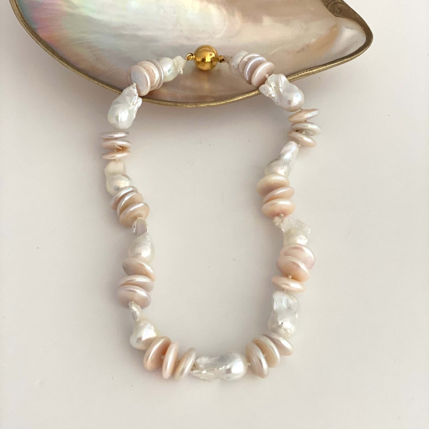A beaded necklace made of irregular baroque pearls in soft white, cream, and pale pink tones, accented with small gold spacer beads, displayed on a light beige surface with a mother-of-pearl shell prop at the top.