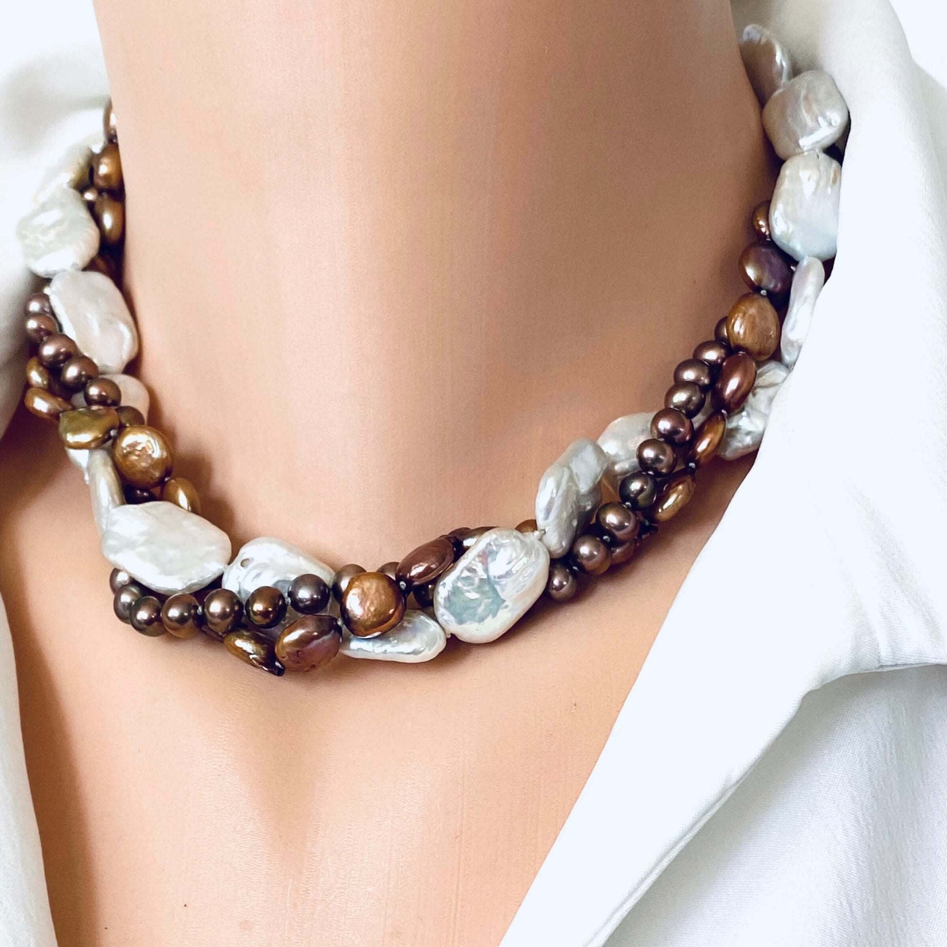 A close-up of a multi-strand statement necklace worn on a mannequin neck. The necklace features large, irregular white baroque pearls, dark brown beads, and metallic bronze beads, styled with a white collared shirt.
