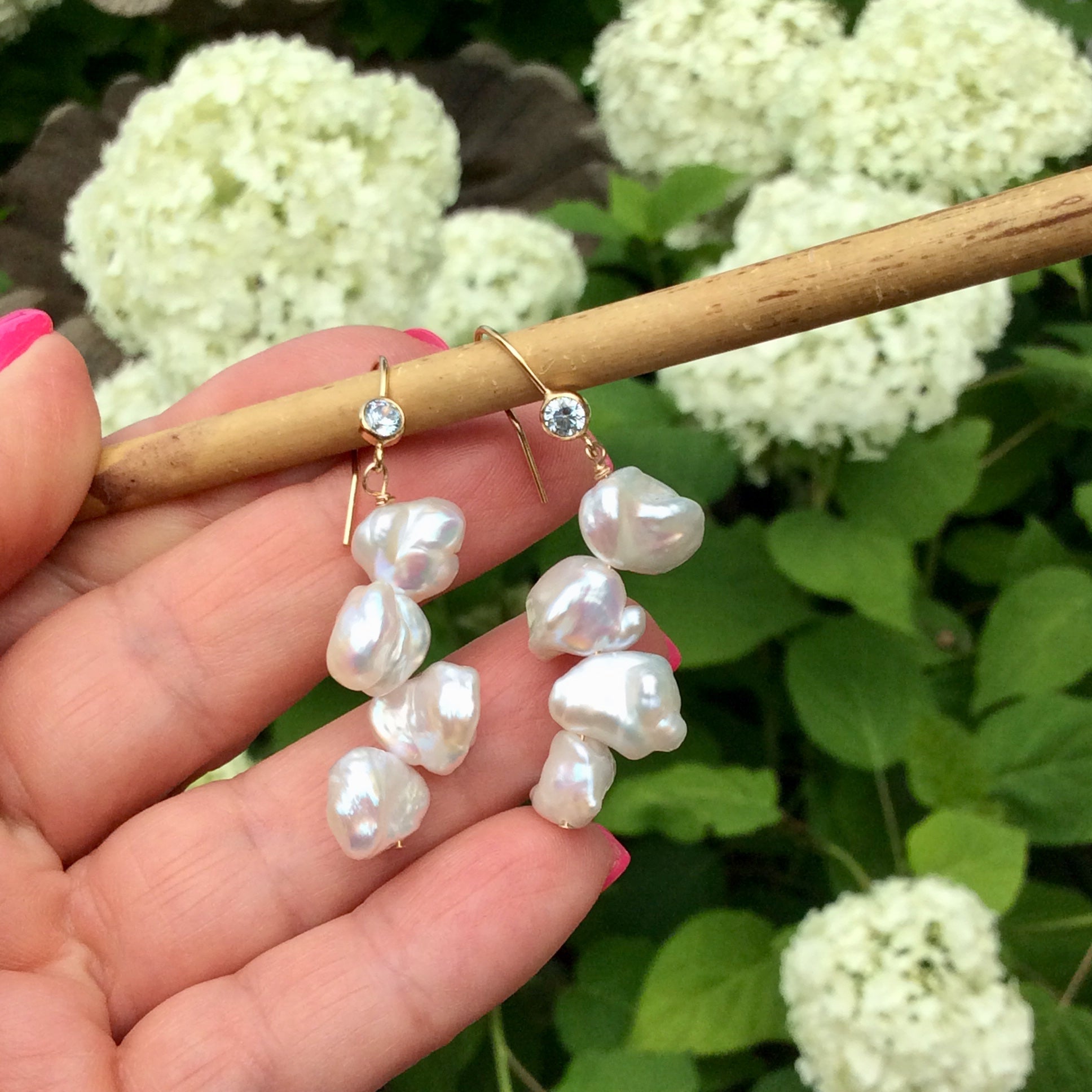 Pearl drop earrings showcasing keshi pearls and blue CZ, held elegantly among lush greenery.