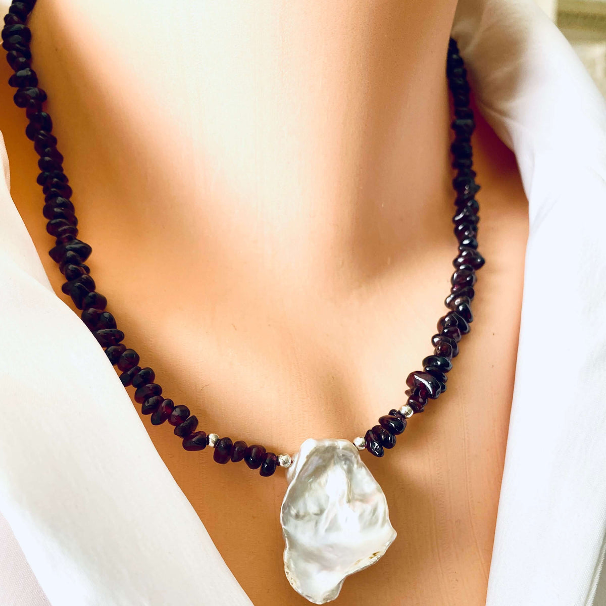 A close-up of a necklace displayed on a mannequin bust wearing a white top. The necklace features a strand of dark, red beads with a large, irregular silver pearlpendant.