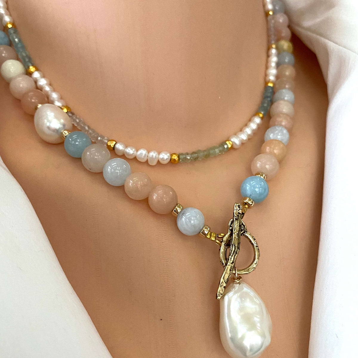 A close-up of a double-strand beaded necklace worn on a model's neck. The necklace features a mix of pastel-colored beads including light blue, peach, and white pearls, accented with small gold spacers and a gold toggle clasp with a large baroque pearl pendant.