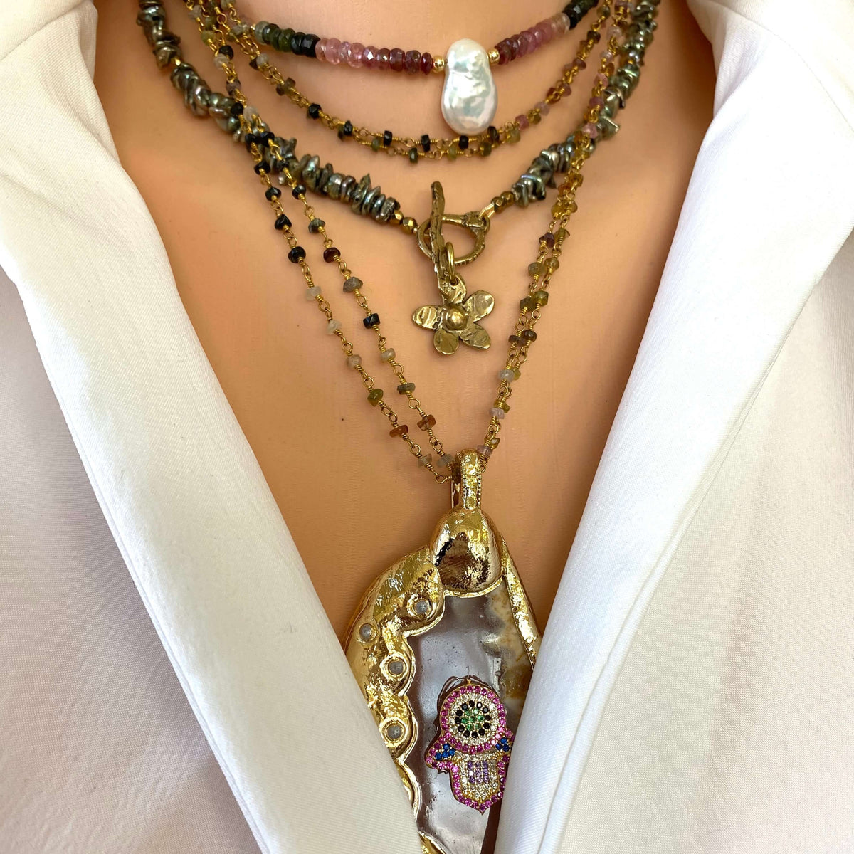 A close-up of a person wearing a layered necklace set with gold chains, beads, and a large ornate pendant featuring a colorful Hamsa hand design over a white shell.